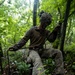 A military service member in MARPAT woodland camouflage uniform navigates a jungle terrain obstacle course using a rope or cable line. The soldier is wearing a tactical helmet with netting and gloves while traversing dense tropical forest vegetation. Additional personnel in similar uniforms are visible in the background, suggesting a group training exercise.