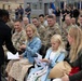 A military ceremony is taking place outdoors, likely a promotion, retirement, or unit recognition event. A female soldier in Army dress uniform is presenting documents or certificates to family members seated in the front row, including two young blonde girls and a woman. Rows of soldiers in Army Combat Uniforms (ACUs) are seated in the background, with additional personnel and flags visible further back.