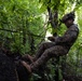 A military service member in digital camouflage uniform is rappelling or rope-traversing down a rocky surface in a dense jungle or forest environment. The soldier is equipped with tactical gear including a helmet with night vision device mount and appears to be managing a rope or line. The lush green vegetation and rocky terrain suggest a tropical or subtropical training environment.