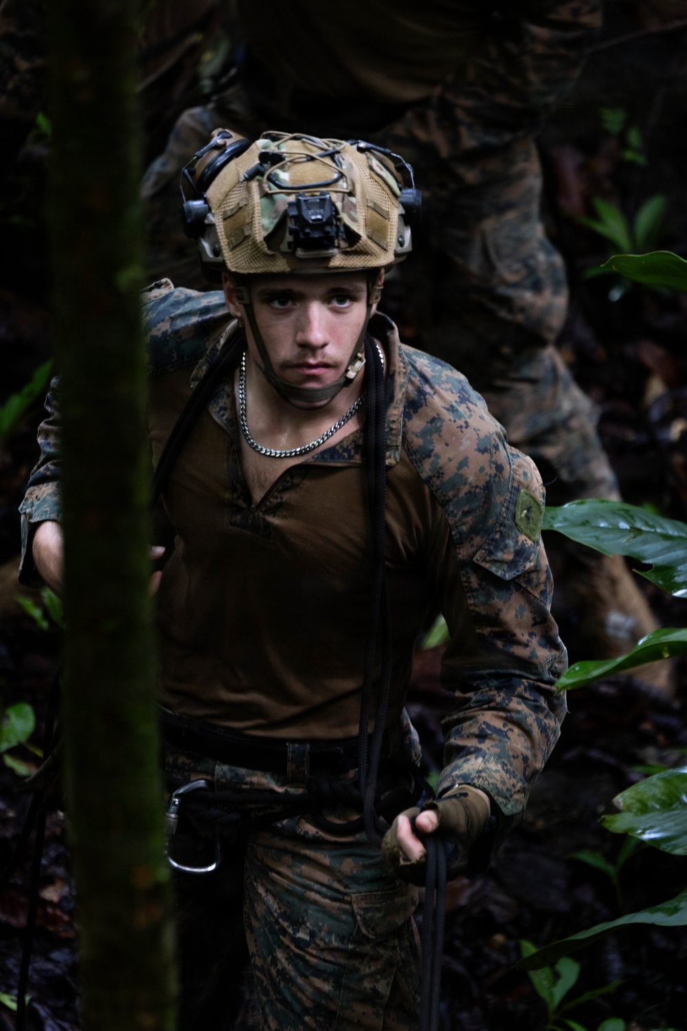 A young male soldier in MARPAT (Marine pattern) camouflage uniform is moving through dense jungle terrain. He is equipped with a tactical helmet fitted with a night-vision goggle mount and is holding a rope or cord, suggesting a rappelling or rope-traversal activity. A second soldier in similar uniform is partially visible in the background.