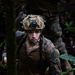 A young male soldier in MARPAT (Marine pattern) camouflage uniform is moving through dense jungle terrain. He is equipped with a tactical helmet fitted with a night-vision goggle mount and is holding a rope or cord, suggesting a rappelling or rope-traversal activity. A second soldier in similar uniform is partially visible in the background.