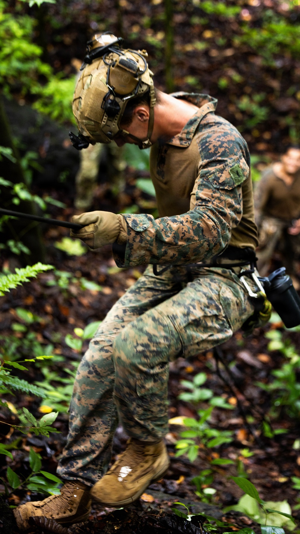 A military service member in MARPAT (Marine pattern) digital camouflage uniform is rappelling or conducting rope work in a dense jungle/forest environment. The soldier is equipped with a tactical helmet fitted with night-vision goggle mounts, gloves, and a rappelling harness with carabiner. Additional personnel are visible in the background, suggesting a unit training exercise.