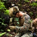 A military service member in MARPAT (Marine pattern) digital camouflage uniform is rappelling or conducting rope work in a dense jungle/forest environment. The soldier is equipped with a tactical helmet fitted with night-vision goggle mounts, gloves, and a rappelling harness with carabiner. Additional personnel are visible in the background, suggesting a unit training exercise.