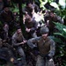 A group of military personnel in camouflage uniforms are navigating through dense jungle terrain, climbing over rocks and vegetation. The foreground soldier wears a tactical helmet with a mounted device and gloves, suggesting a training or operational exercise in a tropical environment. Multiple soldiers are visible in the background, all wearing matching MARPAT (Marine pattern) digital camouflage uniforms.