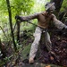 A military service member in camouflage uniform is navigating a steep, rocky, and densely forested terrain while holding onto ropes or cables for support. The individual appears to be conducting a field training exercise or tactical movement through challenging jungle-like terrain. The environment is wet and muddy, suggesting recent rainfall.