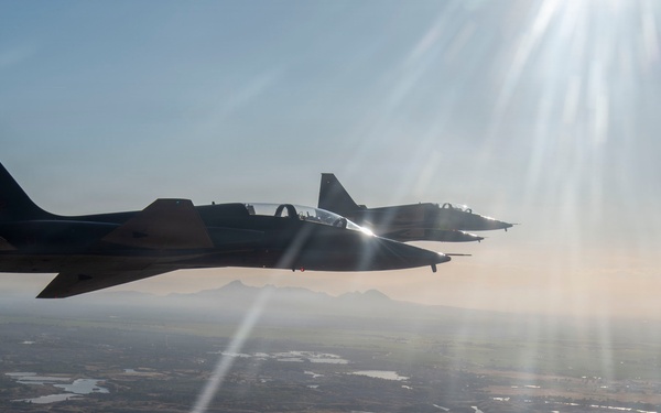 Two dark-colored military jet aircraft are flying in close formation over a flat, semi-arid landscape. The image is taken from a third aircraft flying alongside, capturing the pair against a hazy blue sky with intense sun flare visible in the upper right. The terrain below shows agricultural fields, scattered water bodies, and sparse vegetation.