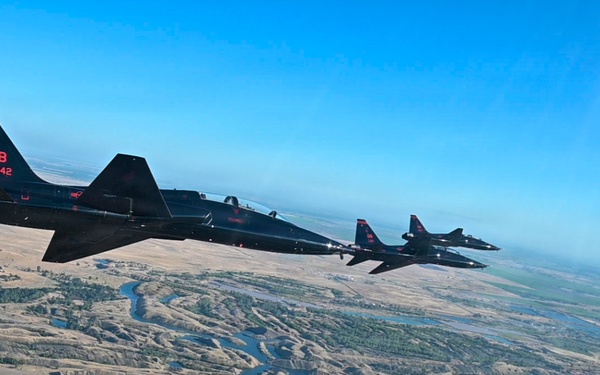 Three black military jet aircraft fly in formation over a rugged, semi-arid landscape featuring winding rivers and eroded terrain. The lead aircraft displays tail markings 'BB' and serial number '342' in red lettering. The image appears to be taken from a fourth aircraft flying alongside the formation.