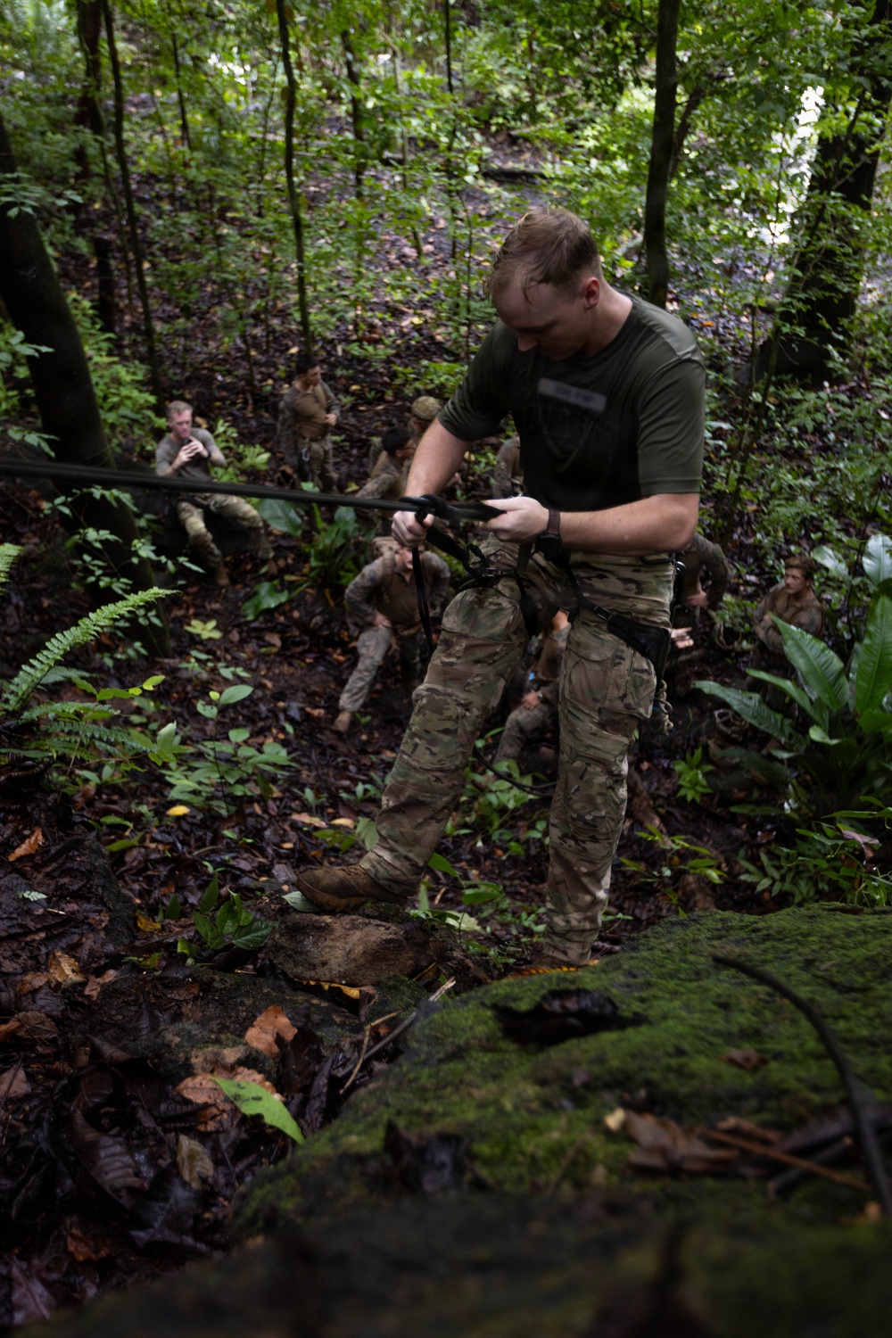 Military personnel are conducting jungle training exercises in a dense tropical forest environment. The foreground subject, wearing multicam camouflage and an olive green t-shirt, is handling a rope or line, likely engaged in a rappelling or rope-crossing exercise. Several other personnel in similar uniforms are visible in the background observing or waiting their turn.