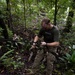 Military personnel are conducting jungle training exercises in a dense tropical forest environment. The foreground subject, wearing multicam camouflage and an olive green t-shirt, is handling a rope or line, likely engaged in a rappelling or rope-crossing exercise. Several other personnel in similar uniforms are visible in the background observing or waiting their turn.
