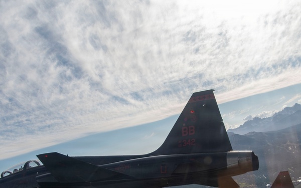 Two dark-painted jet aircraft are flying in formation over a mountainous, forested landscape. The lead aircraft displays tail markings 'BB 342' in red lettering and appears to be a T-38 Talon or similar twin-engine trainer/fighter jet. The image is taken from a third aircraft flying alongside, with sun glare visible in the upper right portion of the frame.