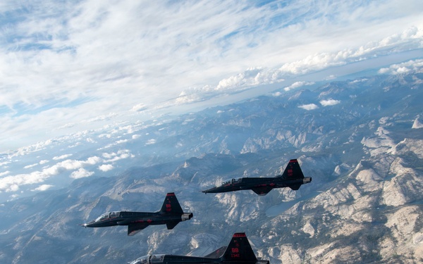 Three dark-colored military jet aircraft fly in a loose formation over a rugged mountainous terrain, likely the Sierra Nevada range in California. The aircraft appear to be T-38 Talon trainer jets with distinctive black paint schemes and red markings. The aerial photograph was taken from another aircraft, capturing both the formation and the dramatic landscape below under a partly cloudy sky.