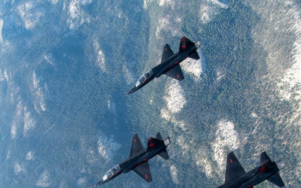 Three dark-colored military jet aircraft are flying in a loose diagonal formation over a mountainous, snow-patched terrain photographed from above. The jets appear to be identical models with dark paint schemes and red markings, consistent with a display or aerobatic team. The aerial perspective provides a dramatic view of both the aircraft and the rugged landscape below.
