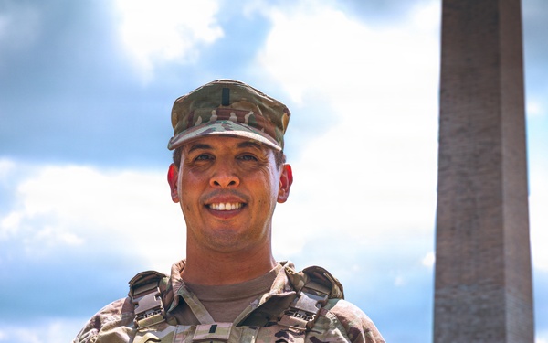 A smiling U.S. Army soldier in full combat uniform and body armor is photographed in front of the Washington Monument in Washington, D.C. The soldier is wearing Operational Camouflage Pattern (OCP) uniform with visible 'ARMY' tape, a name tape beginning with 'ES', and an American flag patch on the right sleeve. The image appears to be an official or semi-official portrait taken on the National Mall.