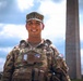 A smiling U.S. Army soldier in full combat uniform and body armor is photographed in front of the Washington Monument in Washington, D.C. The soldier is wearing Operational Camouflage Pattern (OCP) uniform with visible 'ARMY' tape, a name tape beginning with 'ES', and an American flag patch on the right sleeve. The image appears to be an official or semi-official portrait taken on the National Mall.