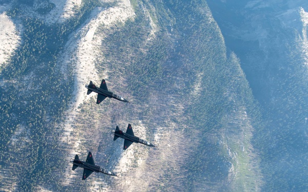 Three military jet aircraft are flying in a diagonal echelon formation over a mountainous, forested terrain. The aircraft appear to be T-38 Talon trainer jets based on their distinctive delta-wing silhouette and twin-engine configuration. The aerial photograph is taken from above and slightly behind, providing a top-down perspective over rugged mountain ridges partially covered with snow and coniferous trees.