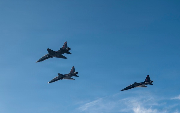 Three military jet aircraft are flying in a loose formation against a blue sky with scattered cirrus clouds. The aircraft appear to be small, twin-engine jets consistent with the Northrop T-38 Talon or F-5 Tiger family, photographed from a low angle looking upward. The formation is staggered diagonally across the frame, suggesting a coordinated flight exercise or aerial demonstration.