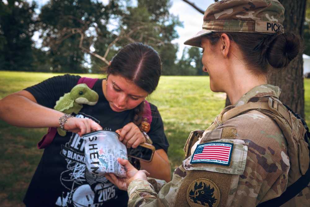 A female soldier in U.S. Army camouflage uniform is interacting with a civilian young woman outdoors in a grassy, tree-lined area. The soldier, whose name tape reads 'PICKL' (partially visible), wears a District of Columbia National Guard patch and is examining or inspecting a clear bag or container held by the civilian. The interaction appears friendly and routine in nature.