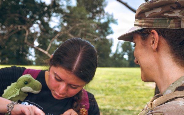 A female soldier in U.S. Army camouflage uniform is interacting with a civilian young woman outdoors in a grassy, tree-lined area. The soldier, whose name tape reads 'PICKL' (partially visible), wears a District of Columbia National Guard patch and is examining or inspecting a clear bag or container held by the civilian. The interaction appears friendly and routine in nature.
