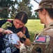 A female soldier in U.S. Army camouflage uniform is interacting with a civilian young woman outdoors in a grassy, tree-lined area. The soldier, whose name tape reads 'PICKL' (partially visible), wears a District of Columbia National Guard patch and is examining or inspecting a clear bag or container held by the civilian. The interaction appears friendly and routine in nature.