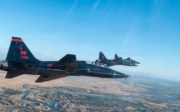 Three black military jet aircraft fly in formation over a rugged, semi-arid landscape featuring a winding river and eroded terrain. The lead aircraft displays tail markings 'BB' and serial number '342' with red insignia. The image is taken from an aerial perspective, likely from a fourth aircraft flying alongside the formation.