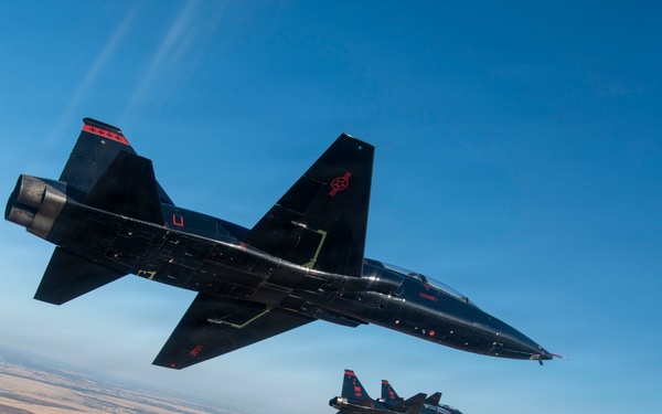 Three black military jet aircraft are flying in formation over a flat, arid landscape with a river system visible below. The lead aircraft is prominently featured in the foreground with two additional aircraft visible in the background. All aircraft feature distinctive black paint schemes with red markings.