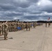 A large formation of U.S. Army soldiers in OCP (Operational Camouflage Pattern) uniforms are standing at attention and rendering a salute on a military airfield tarmac. The ceremony appears to be a formal military event, likely a change of command, deployment, or redeployment ceremony, with guidons and the American flag visible. Military helicopters, identifiable as AH-64 Apaches, are parked in the background.