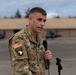 A U.S. Army soldier in OCP (Operational Camouflage Pattern) uniform is speaking into a handheld microphone, likely at an official military event or press briefing. The soldier wears the 101st Airborne Division patch with 'Airborne' tab and a U.S. flag patch on the left sleeve. The setting appears to be an outdoor military installation or airfield.