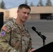 A U.S. Army officer identified by name tape as 'TRIVETTE' stands at a podium with a microphone, appearing to address an audience at an outdoor military installation. He is wearing Operational Camouflage Pattern (OCP) uniform with a 10th Mountain Division patch and U.S. flag on his left sleeve. A black oak leaf cluster rank insignia is visible on his chest, indicating the rank of Lieutenant Colonel.