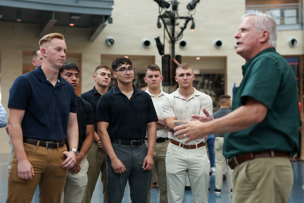 An older man with gray hair wearing a green polo shirt appears to be speaking or presenting to a group of young men in a modern indoor facility. The young men are attentively listening, suggesting a mentorship, orientation, or briefing session. The setting appears to be a contemporary building with visible lighting rigs and architectural features overhead.
