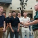 An older man with gray hair wearing a green polo shirt appears to be speaking or presenting to a group of young men in a modern indoor facility. The young men are attentively listening, suggesting a mentorship, orientation, or briefing session. The setting appears to be a contemporary building with visible lighting rigs and architectural features overhead.