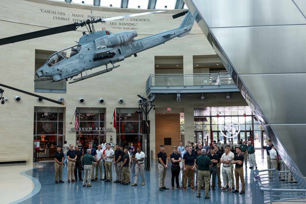 A group of young people, likely students or military recruits, are gathered in the atrium of what appears to be the National Museum of the Marine Corps in Triangle, Virginia. A gray AH-1 Cobra attack helicopter marked 'MARINES' is suspended from the ceiling as a prominent display exhibit. A guide or instructor appears to be addressing the group beneath the aircraft.