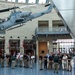 A group of young people, likely students or military recruits, are gathered in the atrium of what appears to be the National Museum of the Marine Corps in Triangle, Virginia. A gray AH-1 Cobra attack helicopter marked 'MARINES' is suspended from the ceiling as a prominent display exhibit. A guide or instructor appears to be addressing the group beneath the aircraft.