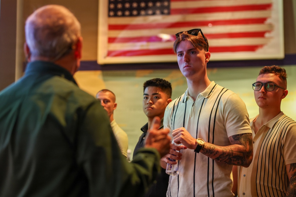 A group of young men are attentively listening to an older man speaking, in what appears to be an indoor venue with an American flag displayed on the wall behind them. The setting suggests a formal or semi-formal gathering, possibly a briefing, orientation, or mentorship event. The young men appear engaged and focused on the speaker.