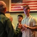 A group of young men are attentively listening to an older man speaking, in what appears to be an indoor venue with an American flag displayed on the wall behind them. The setting suggests a formal or semi-formal gathering, possibly a briefing, orientation, or mentorship event. The young men appear engaged and focused on the speaker.