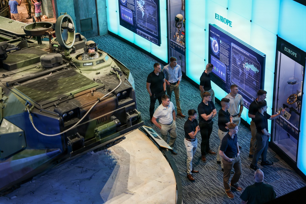 A group of visitors, predominantly young men in casual attire, are touring a military museum exhibit. The exhibit features a large armored military vehicle (appearing to be an AAV or similar amphibious assault vehicle) with a mannequin in the hatch, surrounded by illuminated display panels labeled 'EUROPE'. The scene is photographed from an elevated angle, showing the layout of the exhibit floor.