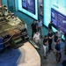 A group of visitors, predominantly young men in casual attire, are touring a military museum exhibit. The exhibit features a large armored military vehicle (appearing to be an AAV or similar amphibious assault vehicle) with a mannequin in the hatch, surrounded by illuminated display panels labeled 'EUROPE'. The scene is photographed from an elevated angle, showing the layout of the exhibit floor.