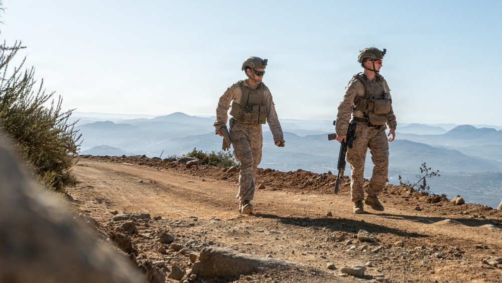 Two military personnel in full combat gear are walking along a dirt ridge road at high elevation. They are wearing matching camouflage uniforms and tactical equipment, with a sweeping mountainous landscape visible in the background. The image appears to depict a patrol or training exercise in an arid, elevated terrain.