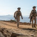 Two military personnel in full combat gear are walking along a dirt ridge road at high elevation. They are wearing matching camouflage uniforms and tactical equipment, with a sweeping mountainous landscape visible in the background. The image appears to depict a patrol or training exercise in an arid, elevated terrain.