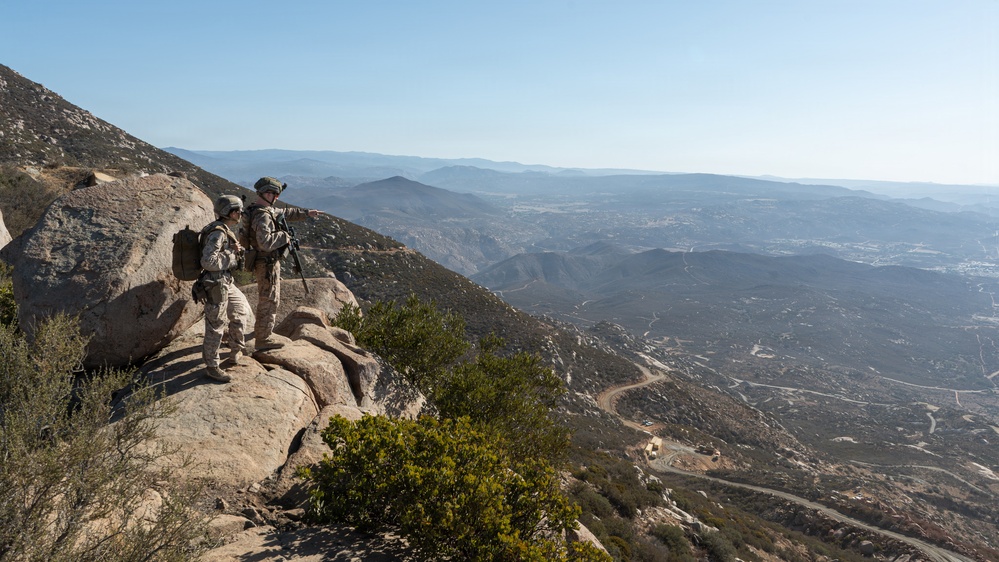 Two military personnel in full combat gear are positioned on a rocky mountain outcrop, observing a wide valley below. One individual appears to be using optical equipment mounted on a tripod while the other points toward the distance. The elevated vantage point provides a sweeping panoramic view of a mountainous landscape with scattered development visible in the valley.
