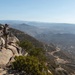 Two military personnel in full combat gear are positioned on a rocky mountain outcrop, observing a wide valley below. One individual appears to be using optical equipment mounted on a tripod while the other points toward the distance. The elevated vantage point provides a sweeping panoramic view of a mountainous landscape with scattered development visible in the valley.