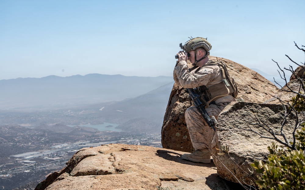 A military soldier in desert camouflage uniform is positioned on a rocky mountain summit, using binoculars to observe a valley below. The soldier is equipped with full combat gear including a helmet, tactical vest, and assault rifle. The elevated vantage point provides a wide panoramic view of a populated valley with visible water features and mountain ranges in the background.