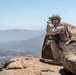 A military soldier in desert camouflage uniform is positioned on a rocky mountain summit, using binoculars to observe a valley below. The soldier is equipped with full combat gear including a helmet, tactical vest, and assault rifle. The elevated vantage point provides a wide panoramic view of a populated valley with visible water features and mountain ranges in the background.