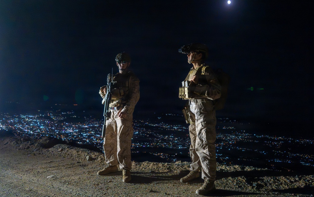 Two armed military personnel in full combat gear stand on an elevated position overlooking a densely lit city at night. Both individuals are wearing camouflage uniforms, tactical vests, helmets with night-vision device mounts, and combat boots. A full moon is visible in the upper right portion of the dark sky.