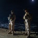 Two armed military personnel in full combat gear stand on an elevated position overlooking a densely lit city at night. Both individuals are wearing camouflage uniforms, tactical vests, helmets with night-vision device mounts, and combat boots. A full moon is visible in the upper right portion of the dark sky.