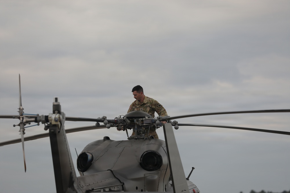 A military service member in camouflage uniform is performing maintenance or inspection work on top of a helicopter. The individual is leaning over the main rotor hub assembly, appearing to inspect or service the rotor head components. The scene takes place outdoors under an overcast sky.