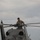 A military service member in camouflage uniform is performing maintenance or inspection work on top of a helicopter. The individual is leaning over the main rotor hub assembly, appearing to inspect or service the rotor head components. The scene takes place outdoors under an overcast sky.