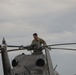 A military service member in camouflage uniform is performing maintenance or inspection work on top of a helicopter. The individual is leaning over the main rotor hub assembly, appearing to inspect or service the rotor head components. The scene takes place outdoors under an overcast sky.
