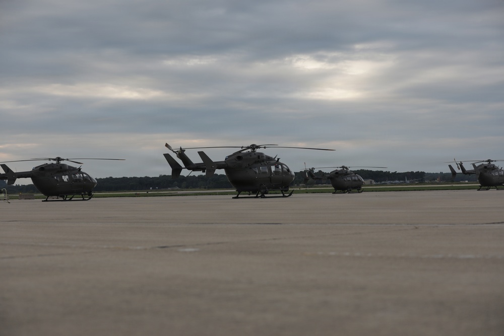 Multiple military helicopters are parked on a concrete flight line or tarmac under an overcast sky. The helicopters appear to be UH-72 Lakota (Eurocopter EC145) utility helicopters in olive drab/dark military paint schemes. The setting appears to be a military airfield with a tree line visible in the background.