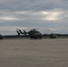 Multiple military helicopters are parked on a concrete flight line or tarmac under an overcast sky. The helicopters appear to be UH-72 Lakota (Eurocopter EC145) utility helicopters in olive drab/dark military paint schemes. The setting appears to be a military airfield with a tree line visible in the background.