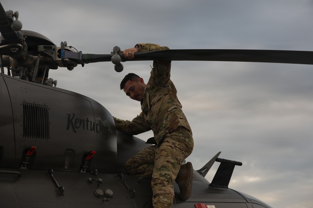 A soldier in MultiCam/OCP camouflage uniform is performing maintenance or inspection on top of a military helicopter. The aircraft has 'Kentucky' written on its fuselage, indicating it belongs to the Kentucky National Guard. The soldier is gripping a main rotor blade while positioned on top of the aircraft under an overcast sky.
